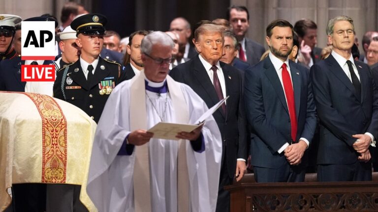 Priest in white robes reads from a booklet at a solemn ceremony, flanked by suited men and a draped casket, with an AP Live logo in the corner.