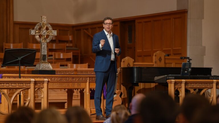 Man in a blue suit delivering a talk on a wood-paneled stage with a decorative cross and piano behind him.
