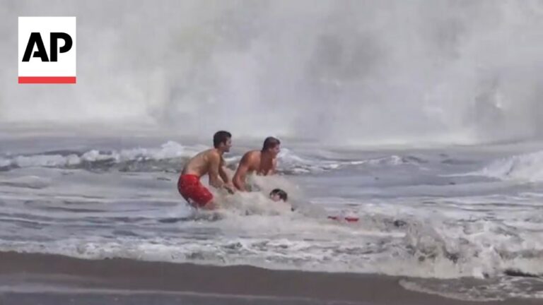 Two shirtless men assist a swimmer in rough surf near the shore; AP logo appears in the upper left corner.
