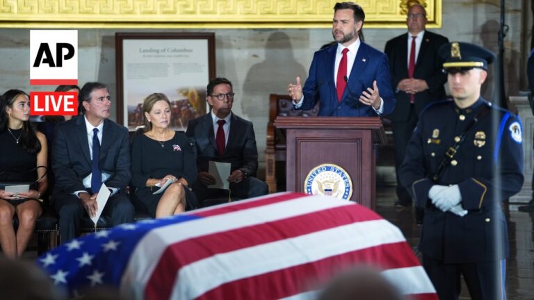 AP Live logo indicates a live news briefing at a formal ceremony with a speaker at a wooden podium bearing the U.S. seal, flanked by seated officials and a uniformed guard nearby. A flag-draped casket is in the foreground.