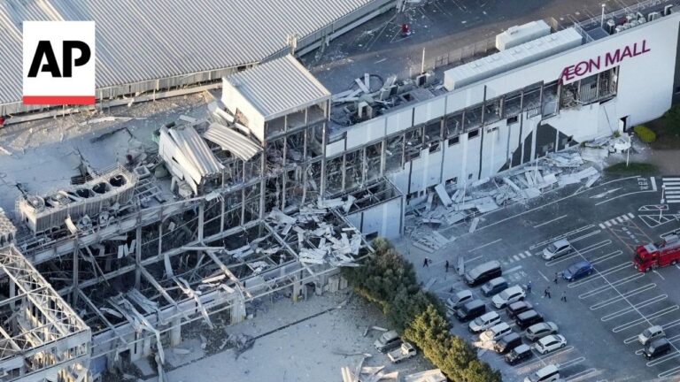 Aerial view of a collapsed mall wing labeled AEON MALL, with rubble and exposed steel beams scattered around a nearby parking lot.