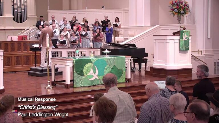 1787516191_maxresdefaultjpg - New Social Gospel Church choir and pianist perform on a raised platform in a church, conductor at left, audience standing in the foreground; green altar cloth and floral arrangement visible; on-screen text reads 'Musical Response', "Christ's Blessing", Paul Leddington Wright.