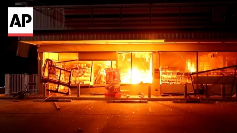 Nighttime storefront fire with bright orange flames, broken windows, and debris across the sidewalk outside a closed shop.