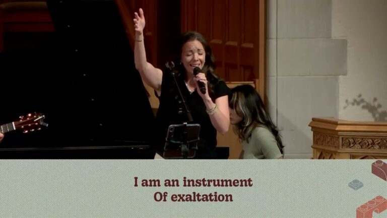 Woman singing into a microphone with her right hand raised on stage, with a banner reading 'I am an instrument Of exaltation' in the foreground.