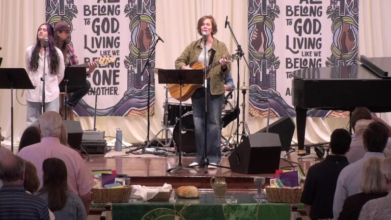 Center-stage singer with an acoustic guitar and microphone, flanked by two backup singers; drummer in back.