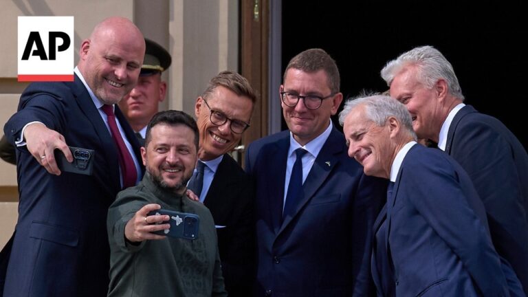 Group of men in suits take a selfie on a doorway step; a man in a green jacket holds the phone, AP logo visible in the corner.