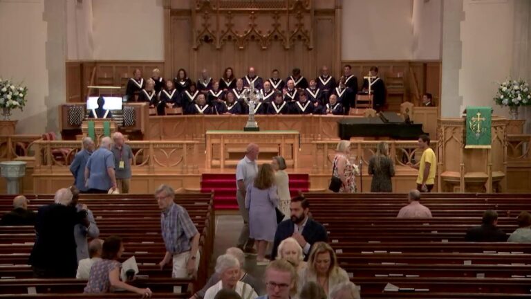 Church sanctuary with a large choir on stage, organ to the left, and a green banner at right; congregants mingle in the pews.