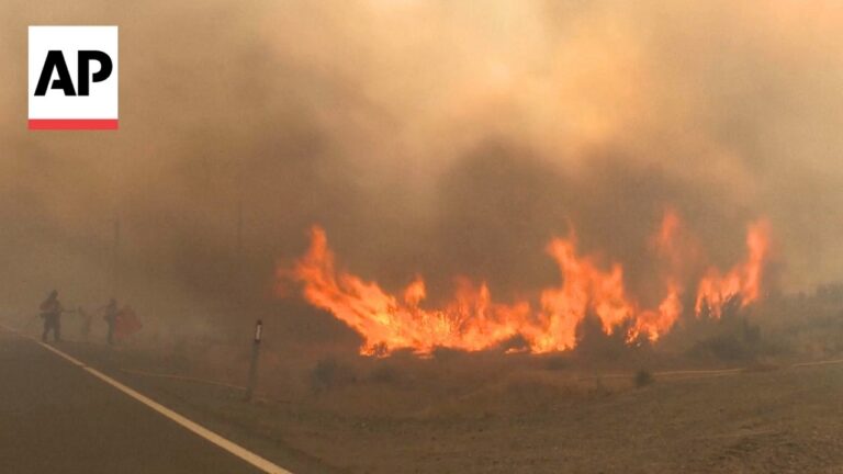 AP logo in the upper left with a wildfire burning along a road in smoky, hazy air behind silhouettes of people and firefighters.