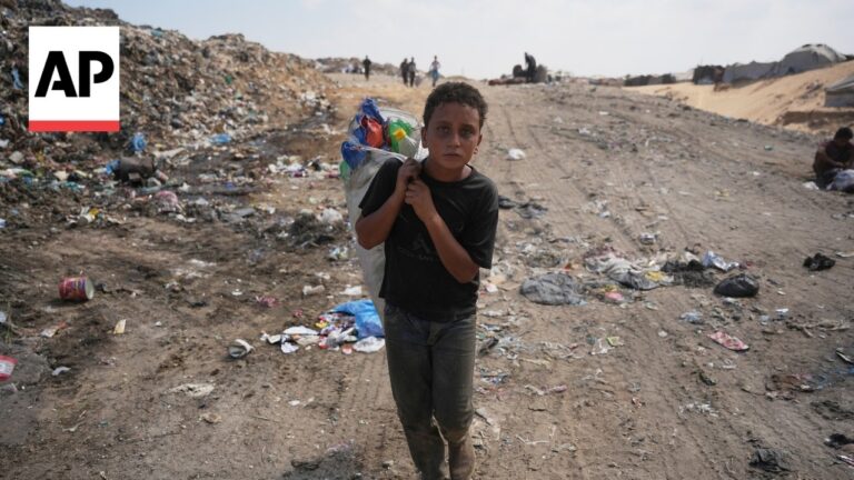 Young boy carrying a bag of belongings walks through a garbage-filled dirt field with tents in the distance; AP logo in the corner.