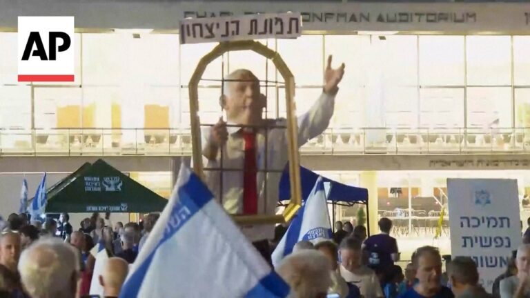 Protester inside a wooden cage raised above the crowd, gesturing with one hand among blue and white Israeli flags and banners outside a building with AP logo nearby.