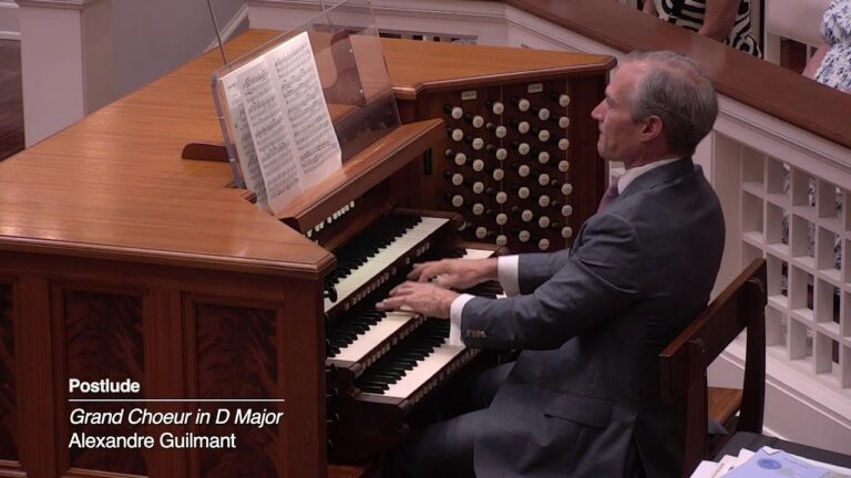 Man in a suit playing a wooden pipe organ at a church, with sheet music on the bench in front of him.