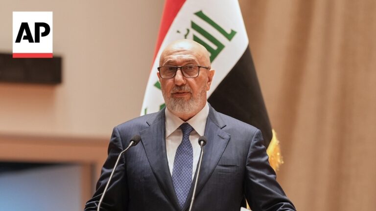 Man in a dark suit and tie speaks at a podium with two microphones, Iraqi flag in the background and AP logo in the top-left corner