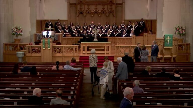 Church interior with a choir in black robes on stage behind the altar, while congregants sit or stand in wooden pews and chat in the aisle.