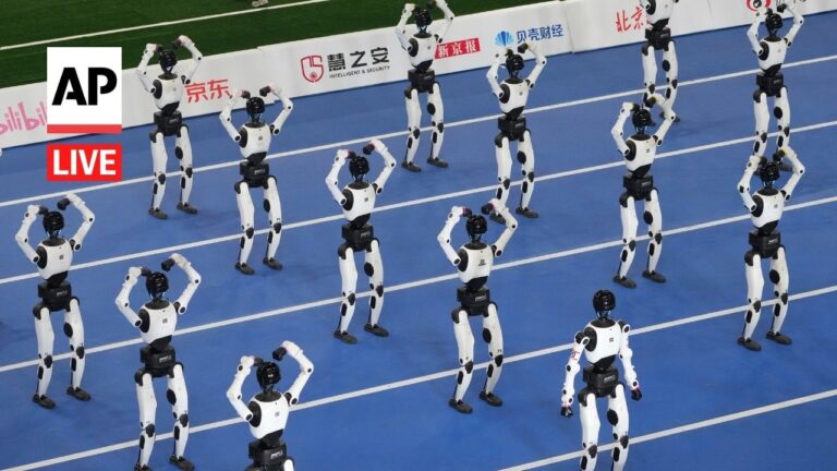 Group of white and black humanoid robots standing on a blue running track, arms raised in synchronized pose, with AP Live logo on the left.
