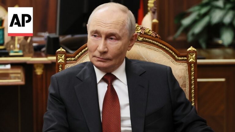 A man in a dark suit and red tie sits in an ornate, carved chair in a formal office with an AP logo in the corner.