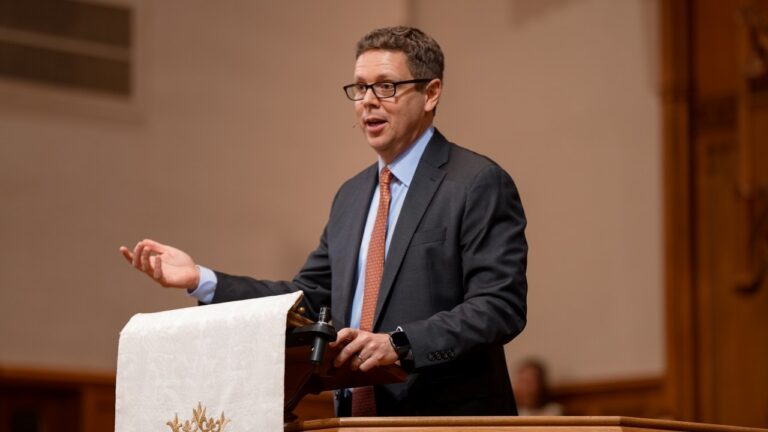 A man in a suit and glasses speaks at a podium, gesturing with his hand during a formal presentation in a conference hall.