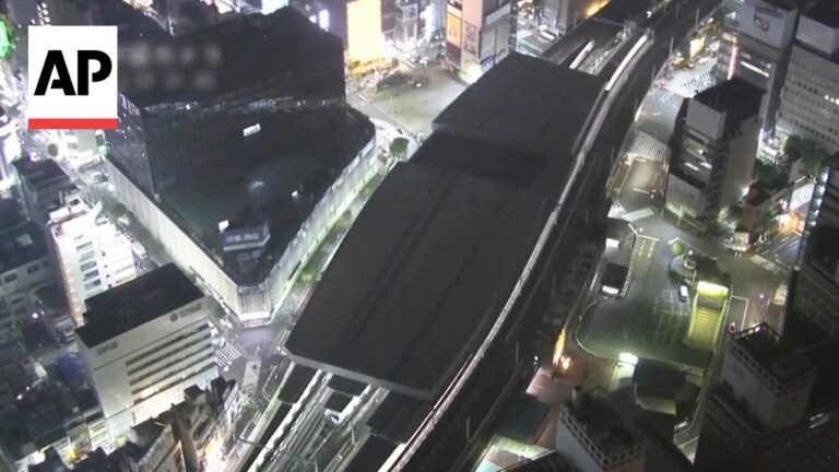 Nighttime cityscape featuring an elevated rail or monorail track above a street, with illuminated buildings and signage; AP logo in the upper-left corner.