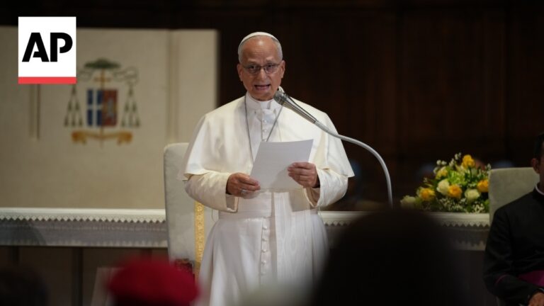 Religious leader in white robes stands at a lectern reading from a sheet, with a microphone and AP logo in the corner.