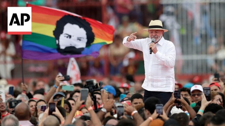 AP photo of a man in a white shirt and straw hat speaking into a microphone at a large rally, with a rainbow flag in the background.