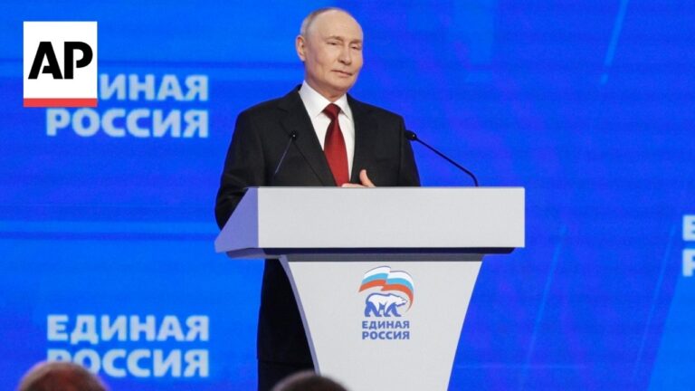 Man in a black suit delivering a speech at a white podium with a blue backdrop displaying the United Russia logo in Cyrillic.