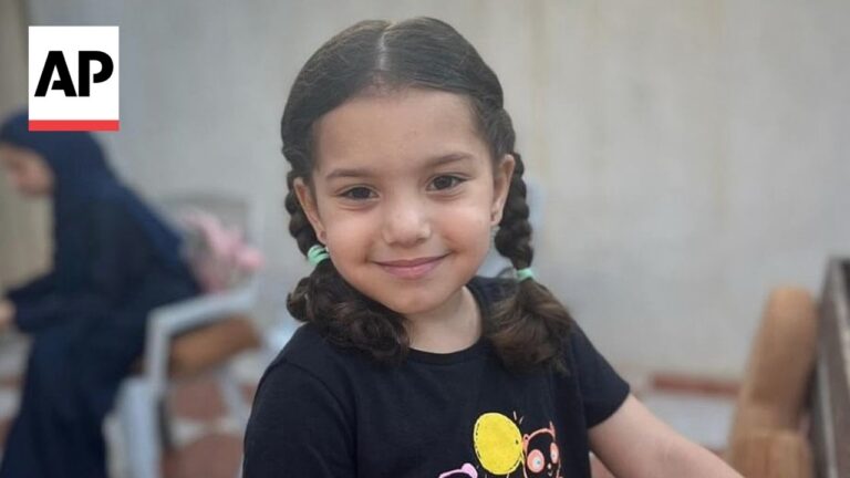 Smiling young girl with two braided pigtails, wearing a dark T-shirt, indoors; AP logo in the top-left corner.