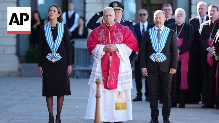 AP photograph showing a central church official in red vestments flanked by two formally dressed aides with blue sashes at a ceremonial event, with clergy in the background.