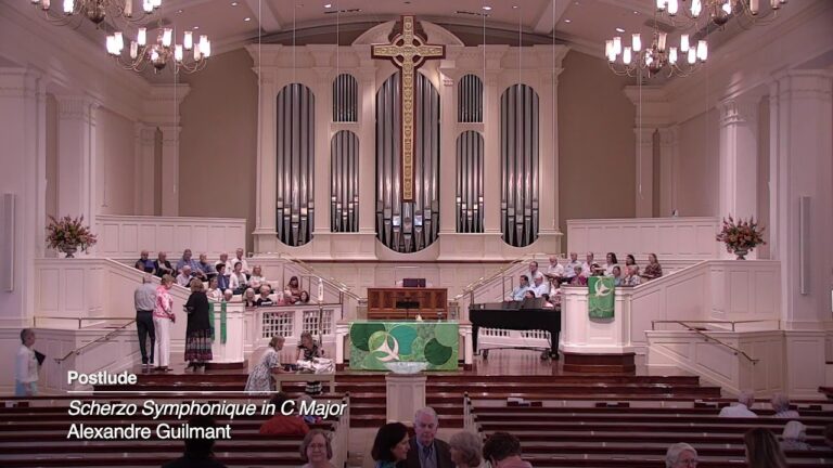 Interior of a church with a large pipe organ and cross at the back, a choir on raised platforms, audience seated in pews, and green decorative banners near the lectern.