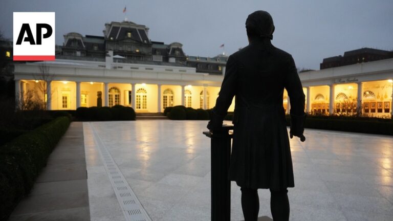 Back view of a tall statue overlooking a lit neoclassical colonnade at dusk; AP logo in the top-left corner.
