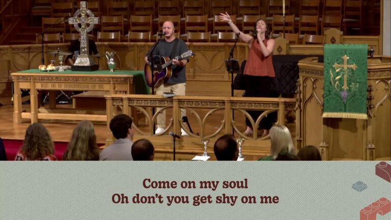 Man with a guitar and a woman singer perform on a church stage, microphone stands and liturgical decor visible behind them.