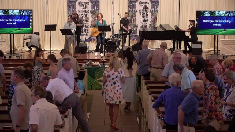 Worship band performing onstage in a church sanctuary while attendees mingle in the pews and walk down the central aisle; two screens display 'Thank you for worshiping with us!'