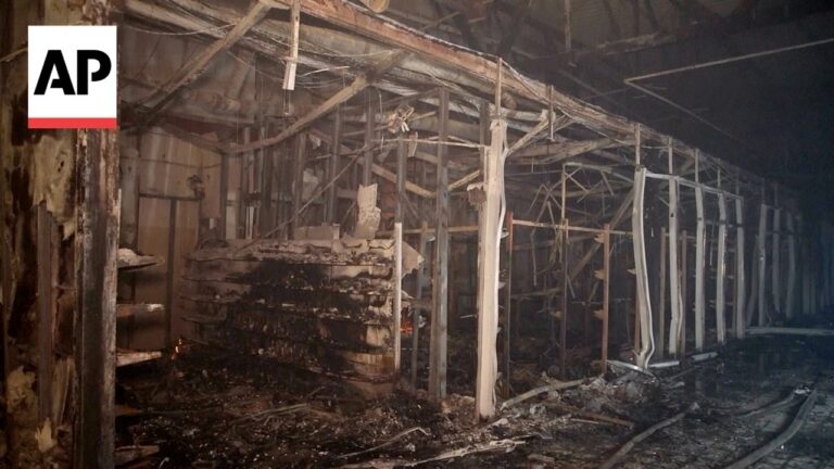 Charred interior of a burned building with collapsed wooden framing and ash-covered debris; AP logo in top-left.