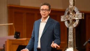 Man in a blue suit and glasses speaks with hands gesturing, standing beside a large ornate cross in a church.|-1