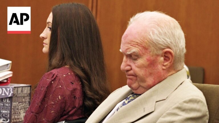 AP logo in the top-left corner; a young woman with long dark hair sits beside an elderly man in a light suit in a courtroom, with books nearby.