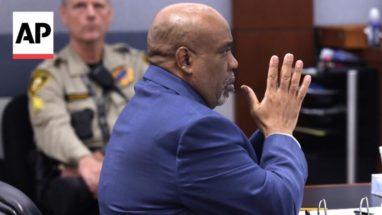 Man in a blue suit sits in a courtroom with hands pressed together, AP logo visible in the top-left corner.