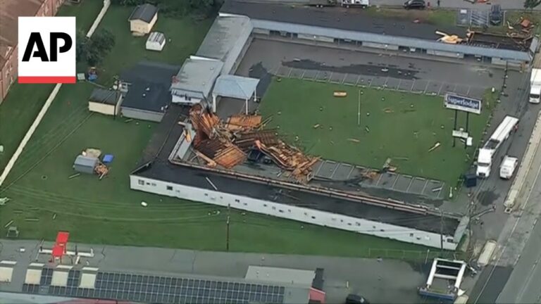 Aerial view of a commercial building with a collapsed roof and scattered wooden debris on the roof and surrounding green lawn. AP logo in the corner.
