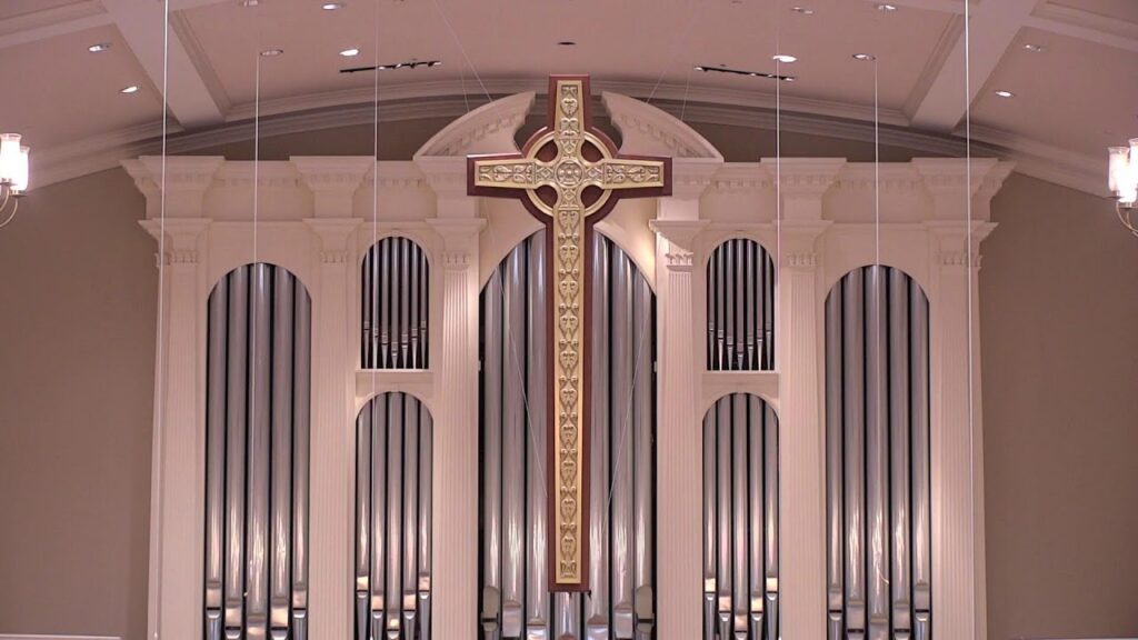 1789286842_maxresdefaultjpg - New Social Gospel Inside a church sanctuary, a large gold cross centerpiece hangs in front of tall organ pipes behind.