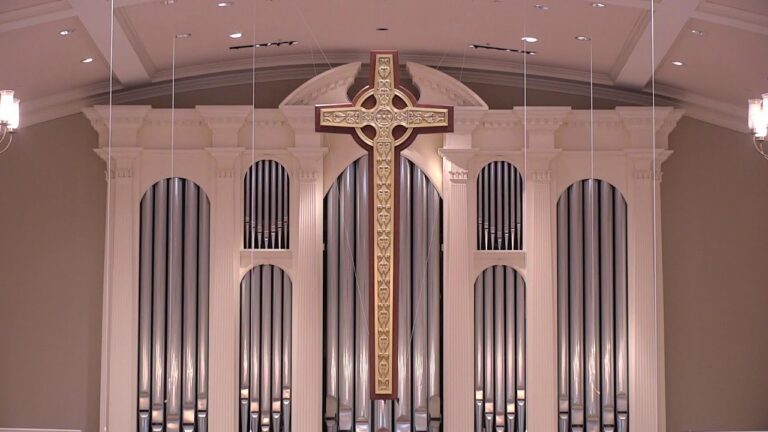 Inside a church sanctuary, a large gold cross centerpiece hangs in front of tall organ pipes behind.