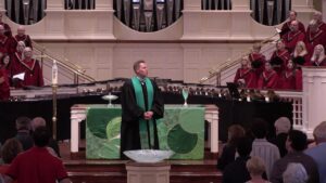 Minister in black robe with green stole stands at pulpit during a church service, choir in red robes behind, congregants seated and standing nearby.