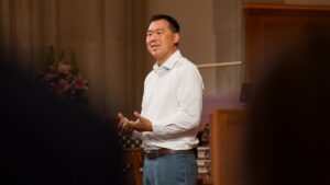 Speaker in a white shirt and blue pants delivers a talk on a stage, gesturing with his hands.