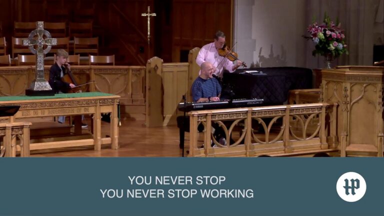 Church musicians performing on a wooden stage: keyboardist singing into a mic with two violinists nearby, ornate woodwork and a cross in the background on stage.