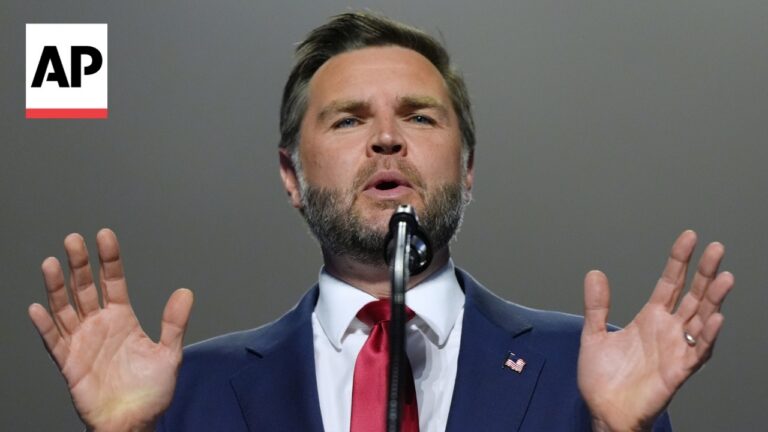 Man in a navy suit and red tie speaks at a microphone, hands raised, AP logo in the corner, American flag pin on lapel.