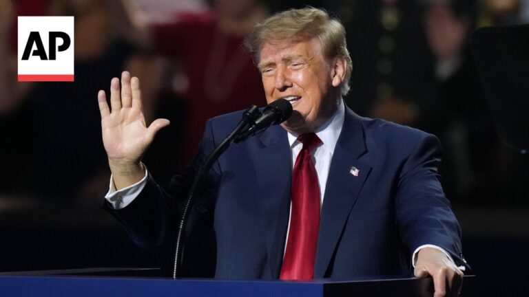 Man in a navy suit and red tie speaks at a podium, raising his left hand; AP logo in the top-left corner.