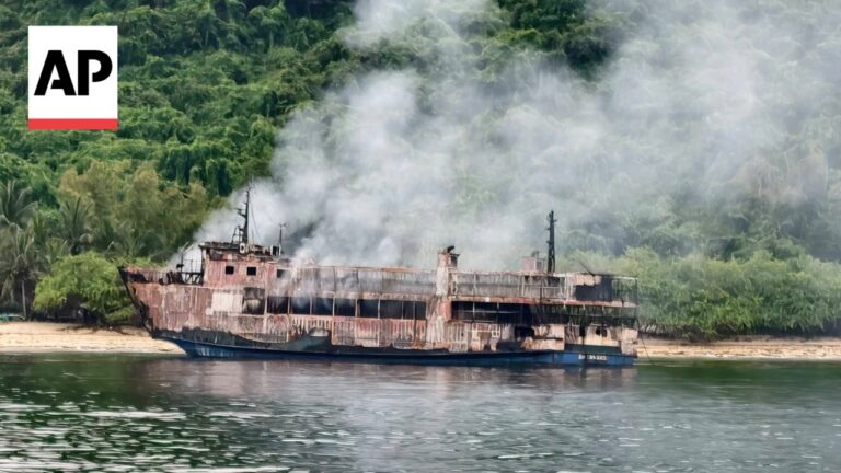 AP news photo of a rusted, burning boat emitting smoke near a tropical shoreline surrounded by dense green foliage