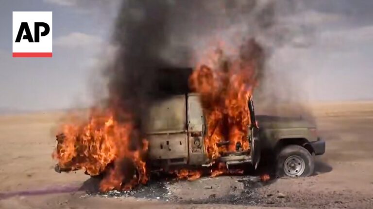 Beige SUV engulfed in large orange flames with black smoke rising in a desert landscape; AP watermark in the top-left corner.