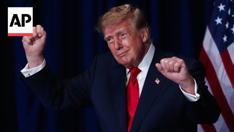 Politician on stage with fists raised, wearing a dark suit and red tie, American flag in the background with an AP watermark.