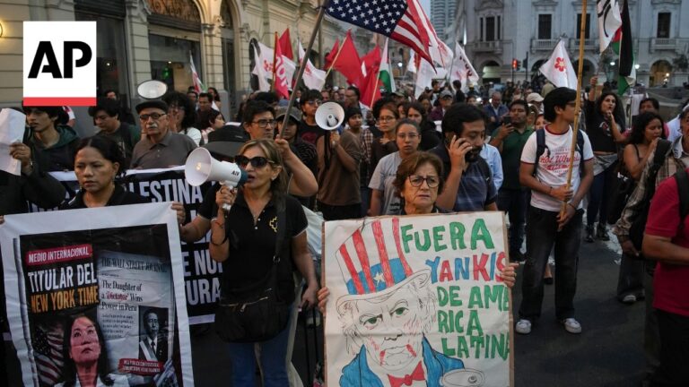 Crowd of diverse protesters marching on a city street, holding banners and a caricature sign saying 'Fuera Yank(s) de America Latina' with an AP logo visible on the left.