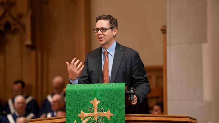 Man in a suit and glasses speaks from a wooden lectern, gesturing with his left hand, in front of a green liturgical banner with a gold cross.