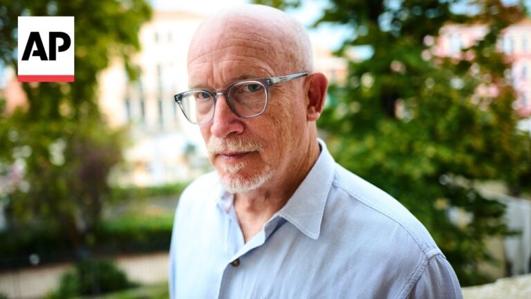 Portrait of an older man with glasses outdoors, wearing a light blue shirt; AP logo visible in the top-left corner.