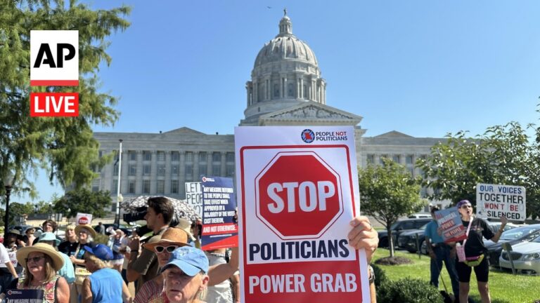 Protest outside a capitol building with a large STOP POLITICIANS sign reading 'POWER GRAB' and an AP Live logo nearby.