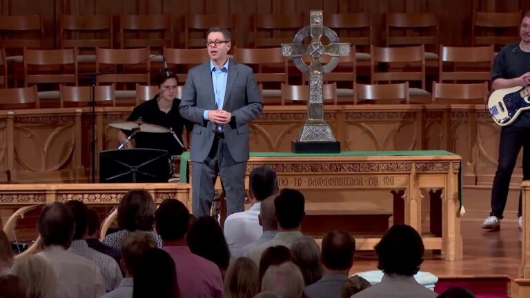Man in a gray suit speaks at a church podium with a large decorative cross, while a drummer and bassist perform in the background and the audience watches.